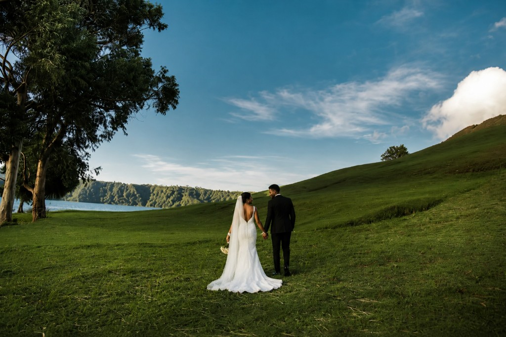 The couple walking hand in hand across a green hillside beside a lake